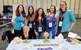 Five smiling people stand behind a table displaying brain models, flyers, and science-themed items at an indoor event.