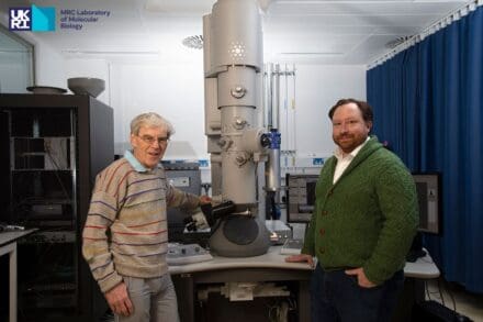Two men, one in a striped grey jumper and the other in a green cardigan, stood either side of scientific equipment