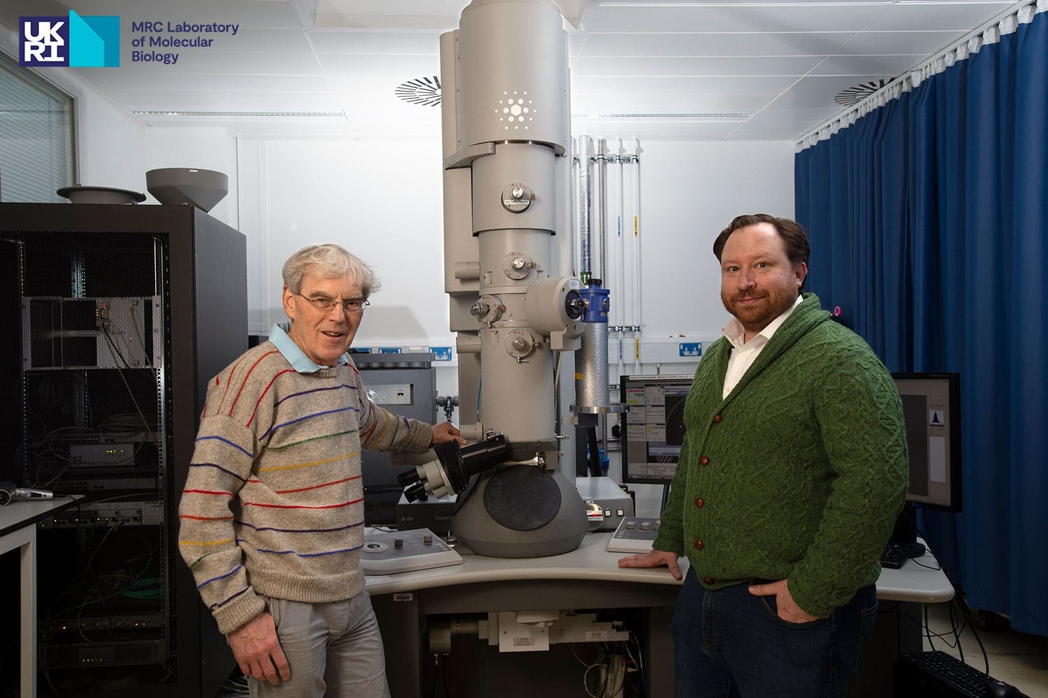 Two men, one in a striped grey jumper and the other in a green cardigan, stood either side of scientific equipment