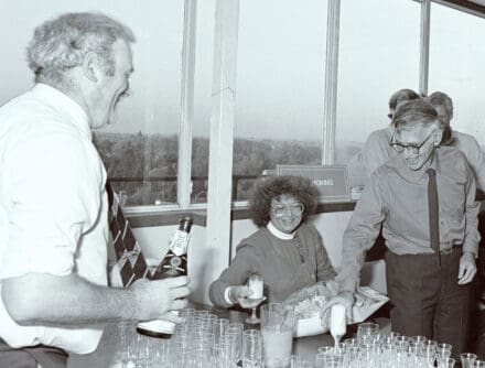 Mike Fuller holding a champagne bottle with many glasses waiting to be filled. Across the table, Liebe Klug holds a glass of champagne and Aaron Klug reaches for a glass which is spilling over