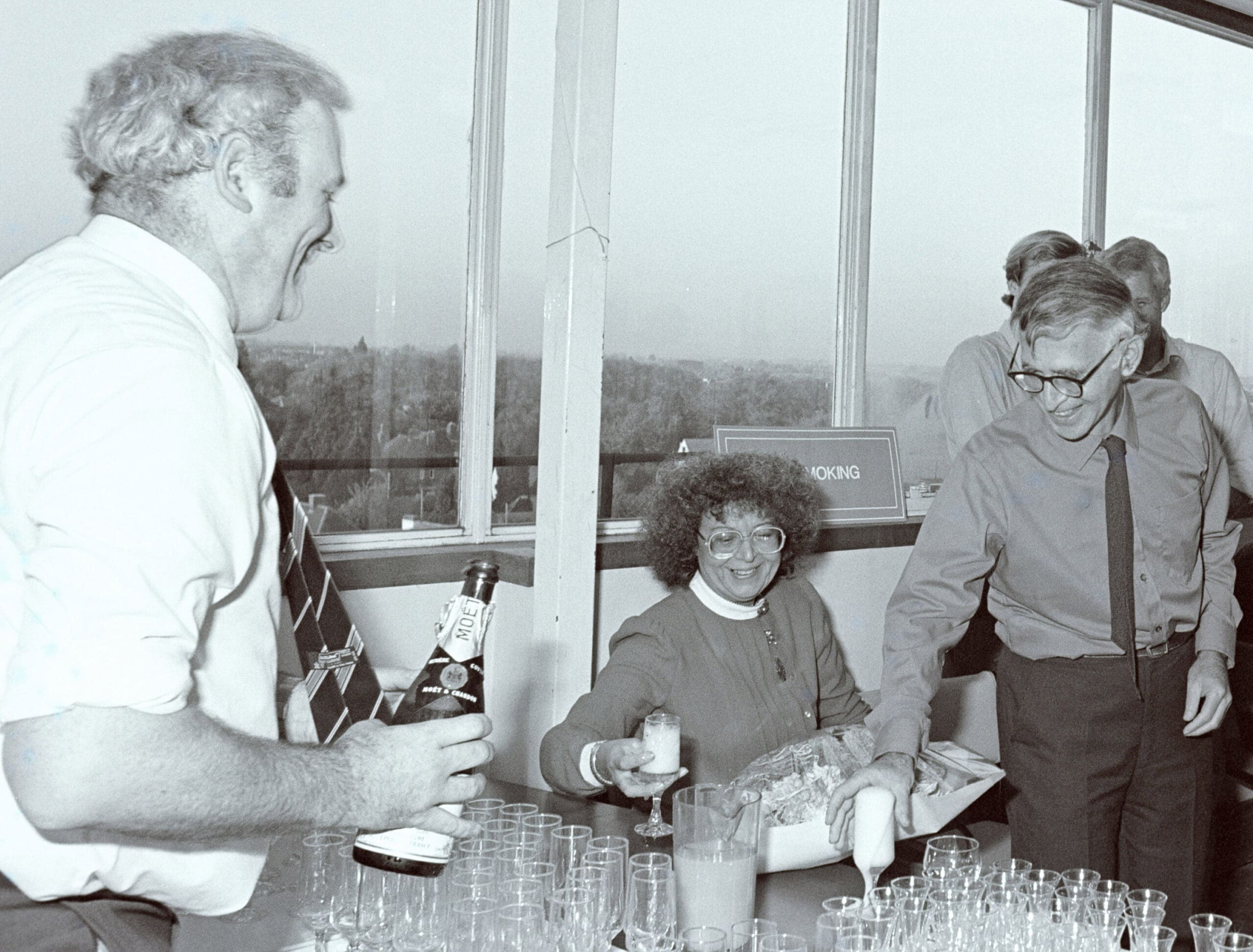 Mike Fuller holding a champagne bottle with many glasses waiting to be filled. Across the table, Liebe Klug holds a glass of champagne and Aaron Klug reaches for a glass which is spilling over