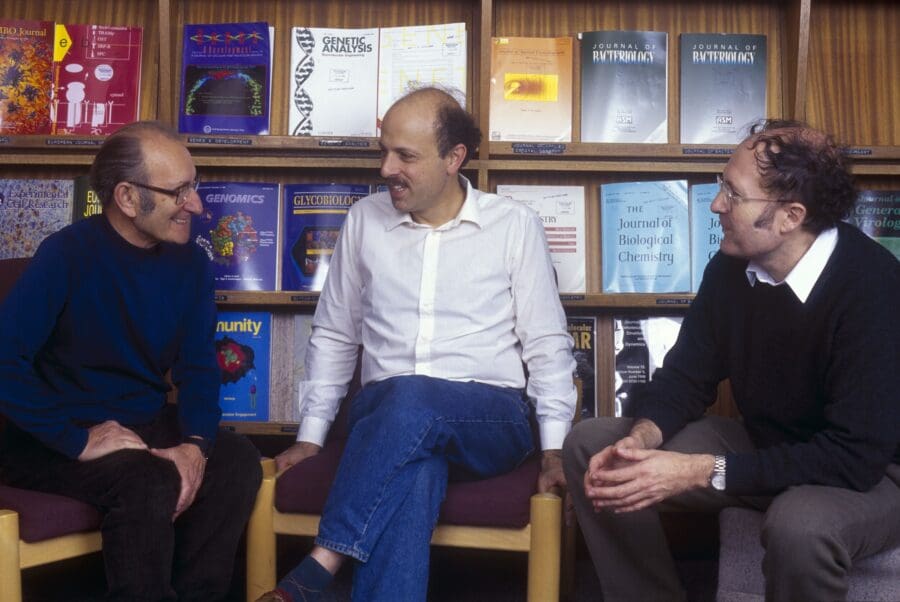 César Milstein, Michael Neuberger and Greg Winter sat on chairs in the LMB Library, behind them, shelves display scientific journals
