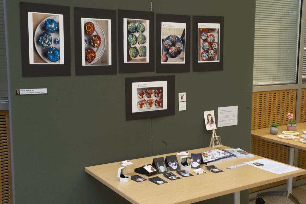 Table against a display board, showcasing jewellery and photographs of decorated cupcakes.