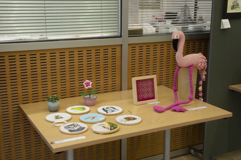 Table displaying embroidery in circular and a square frame, embriodered plants, and a crocheted flamingo.