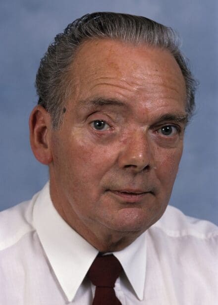 A headshot of a man with grey hair and wearing a white shirt and red necktie
