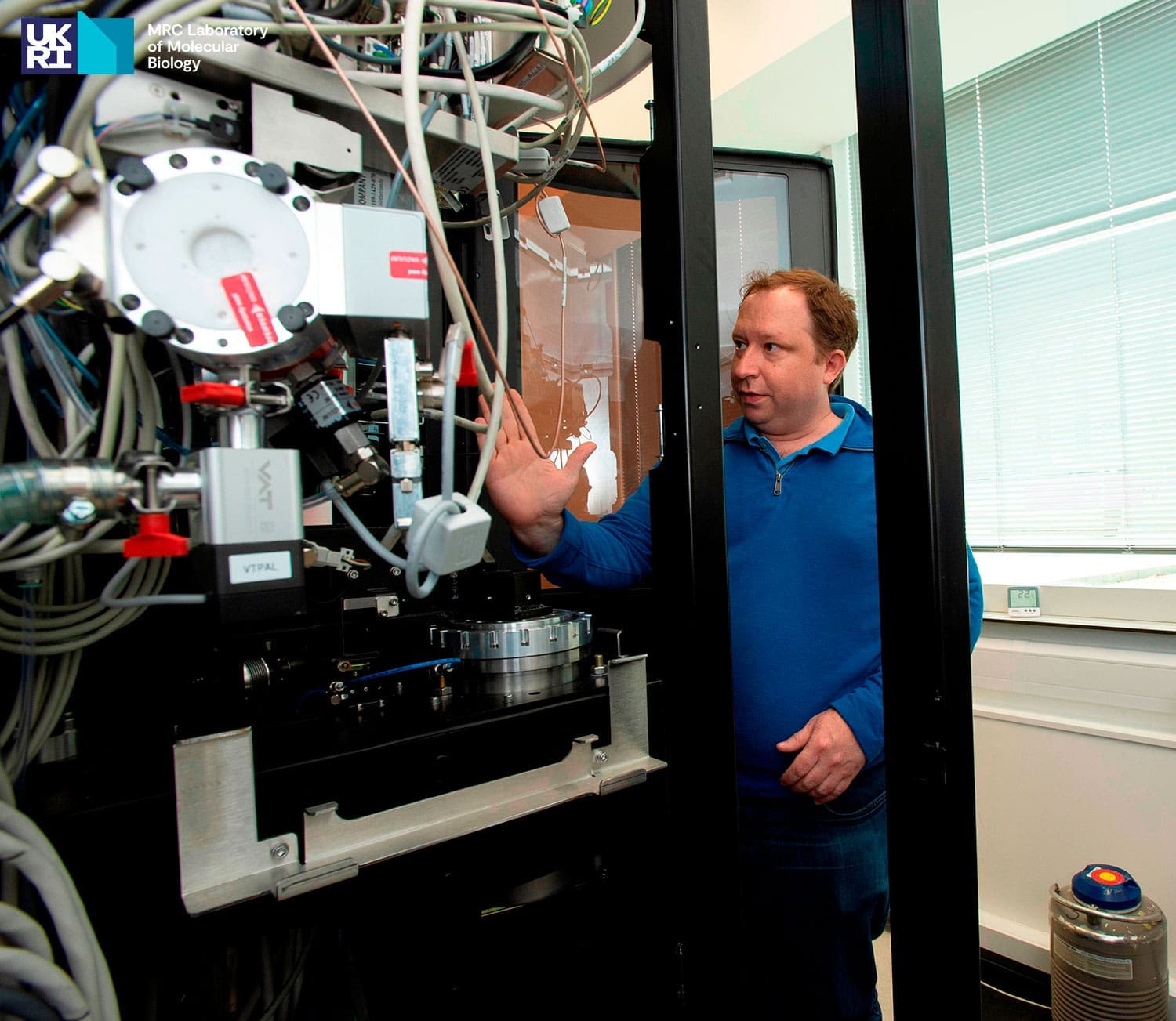 Man in blue sweater looking and pointing to scientific equipment in the foreground with visible wires