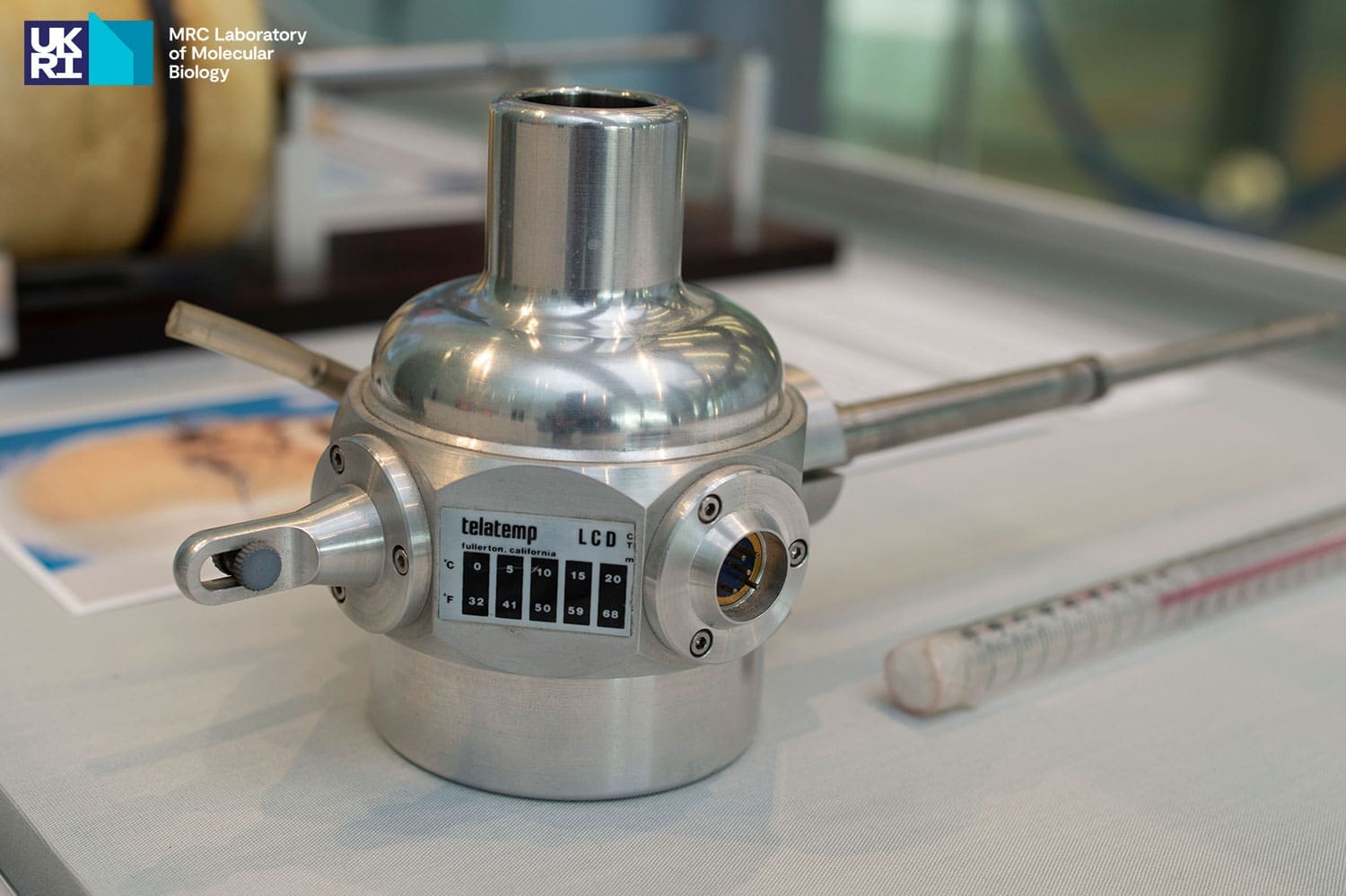 Silver-coloured instrument with a thermometer placed beside it and a colour photographic portrait placed on the other side of it