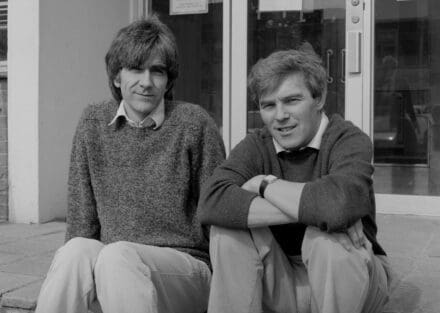 Black and white photo of two men sitting on step outside of glass doors
