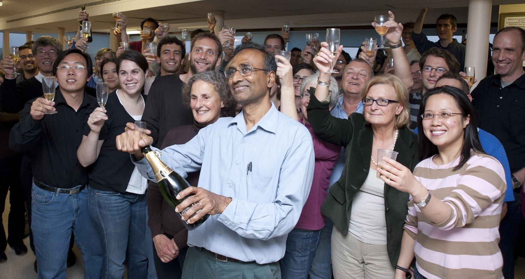 Venki Ramakrishnan opening a bottle of champagne, stood in front of a large crowd of people holding glasses aloft