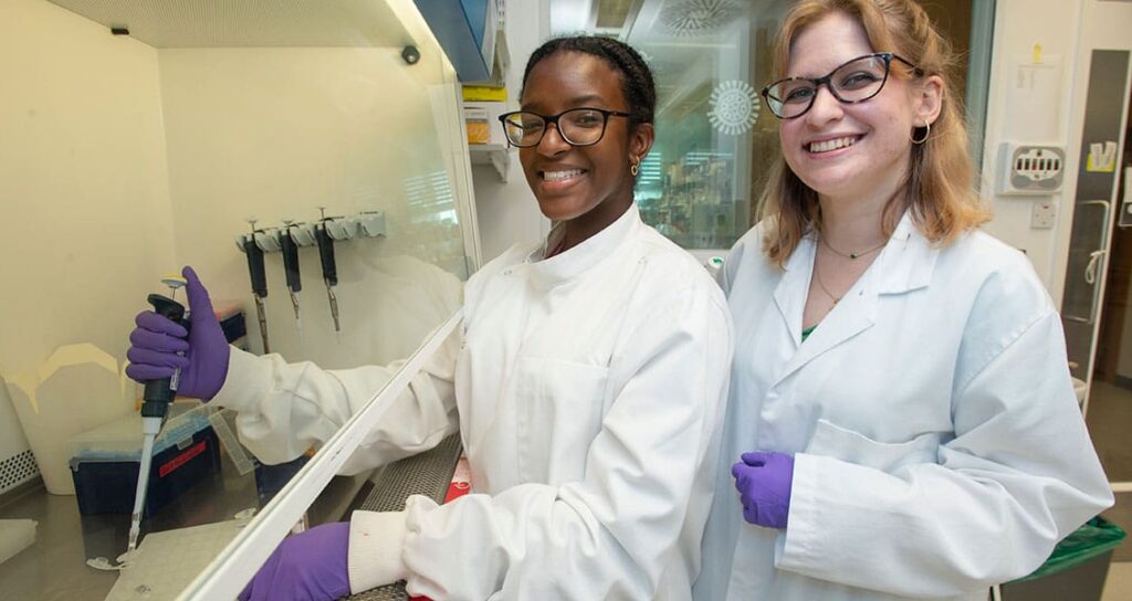 Two smiling young women in lab coats and purple gloves, one using a pipette in a lab hood.