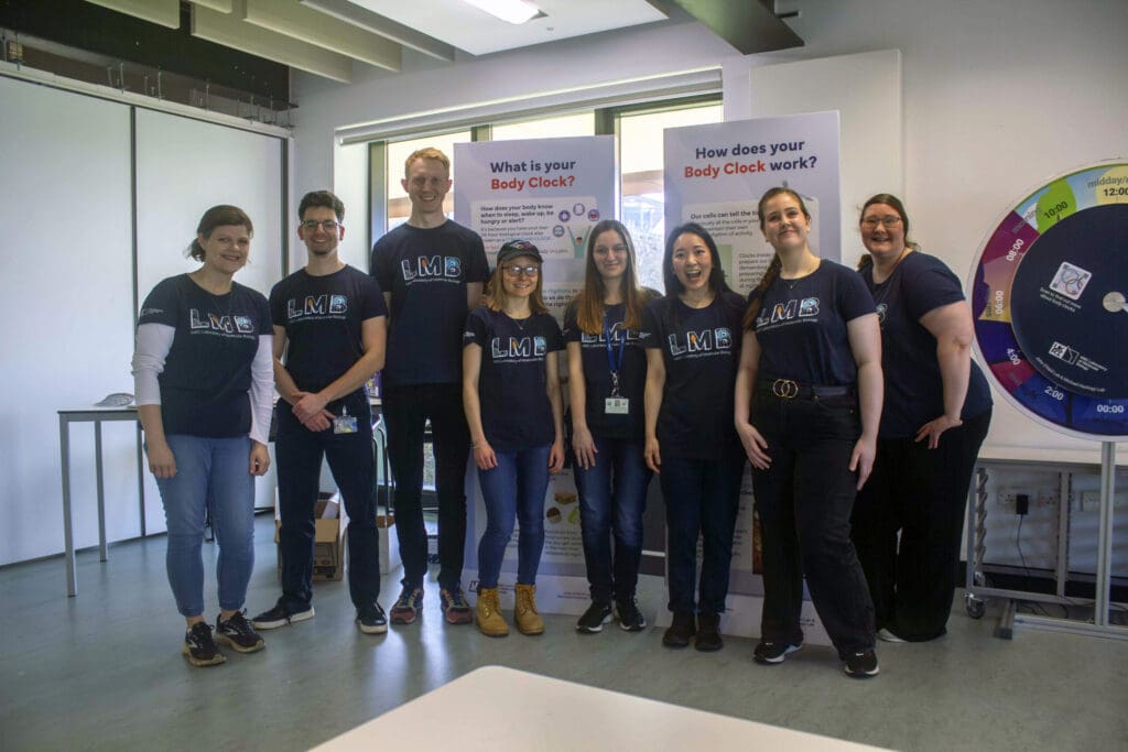 Eight people wearing LMB t-shirts stand smiling in a line, with 'Body Clock' posters and a circular clock display behind them.