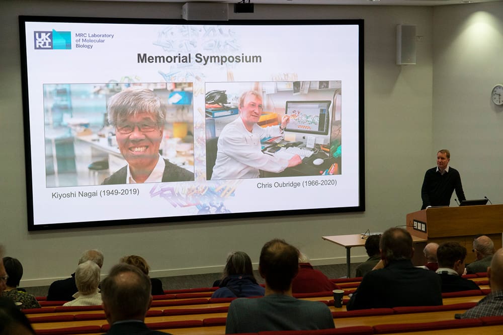 Photo of Jan Loewe giving welcome presentation in the LMB Max Perutz lecture theatre with photos of Kiyoshi and Chris in lab settings on the projector screen.
