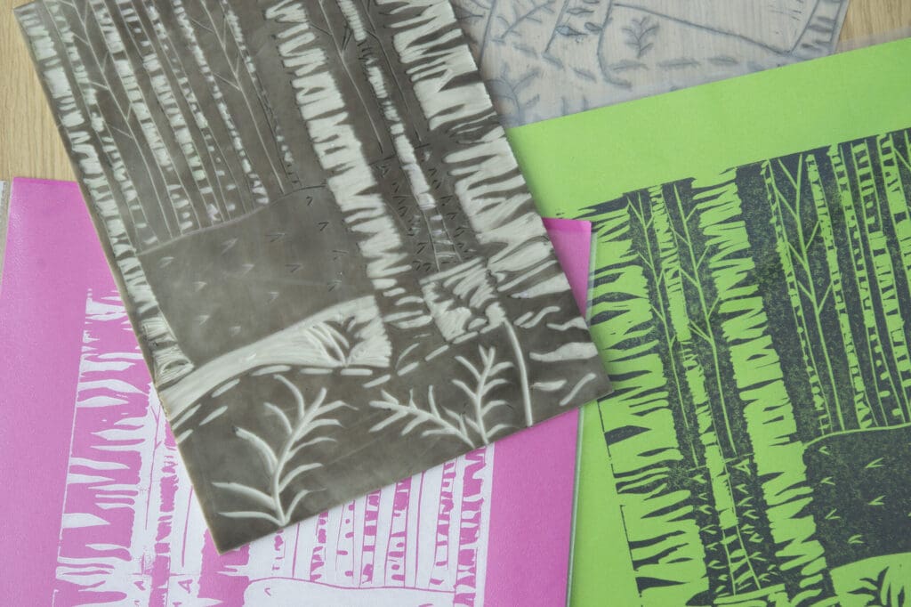 A close up of a printing plate of a forested scene, with black ink on the raised surface. Beneath the printing block are examples of the print, one on pink paper in white ink and the other on green paper in black ink. 