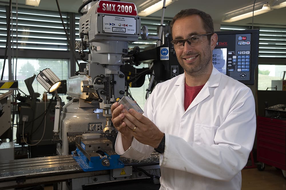 Adam Fowle, wearing a white lab coat stands in front of machining equipment in the LMB’s Mechanical Workshop