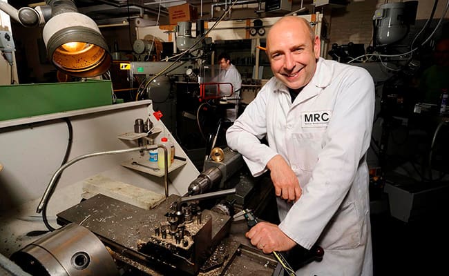 Man wearing lab coat stood next to machinery.